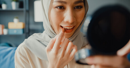 Attractive cheerful young Asian muslim beauty woman in hijab with casual sitting on couch enjoy applying make up lipstick on her lips in cozy living room at home. Girl Islam beauty lifestyle concept.