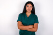 Self confident serious calm Doctor hispanic woman wearing surgeon uniform over white wall stands with arms folded. Shows professional vibe stands in assertive pose.