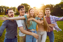 Group Of Happy And Carefree Young Friends Who Are Laughing In Summer Park With Open Arms To Camera. Funny Joyful Young People Having Fun Together Outdoors On Summer Sunny Day. Friendship Concept.
