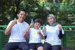 © Gatot - Happy Muslim family holding bottle with water while resting after sport workout outdoors in the public park. Parents and son smiling and giving thumbs up gesture.
