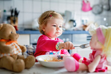 Adorable Baby Girl Eating From Fork Vegetables And Pasta. Food, Child, Feeding And Development Concept. Cute Toddler, Daughter With Spoon Sitting In Highchair And Learning To Eat By Itself.
