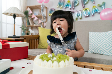 hungry asian toddler baby holding cake knife and enjoying delicious cream while celebrating her second birthday at home decorated for holiday celebration