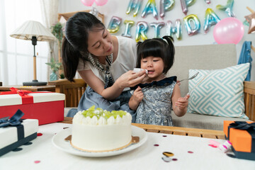 Wall Mural - cute asian baby girl looking at the delicious cake on table with knife in hand while her mother is wiping up cream on her mouth. birthday celebration fun at home