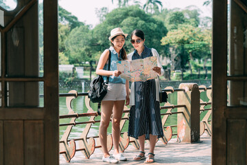 full length view from wooden door cheerful tourist girls are holding and reading a paper map together by the railing near a lake with outdoor sunlight in summer