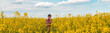 © Bits and Splits - Female farm worker wearing red plaid shirt and trucker's hat standing in cultivated rapeseed field in bloom and looking over crops