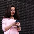 © Granmedia - Beautiful business woman in glasses, in a pink suit, stands against the background of a black brick wall. With phone in hand, typing a message
