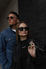 Wall Mural - Fashionable beautiful couple in stylish street fashion outfit with jacket, bag and sunglasses walking on the street on a dark background. Woman's and man's urban casual outwear