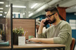 © pressmaster - Young serious employee in eyeglasses and casualwear talking to client on mobile phone while sitting by workplace and looking at laptop screen