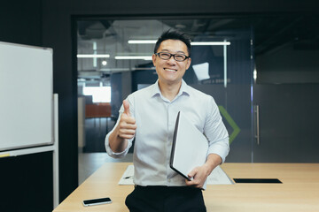 Portrait of successful asian teacher, man in modern classroom, looking at camera and smiling, wearing glasses holding laptop, showing thumbs up gesture affirmative