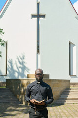 Wall Mural - Young serious priest with open Bible evangelizing audience or telling sermon while standing in front of building of Catholic church