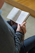 © pressmaster - Hands of senior man with open Bible or Gospel and rosary beads reading verses while sitting in church during sermon or service