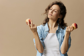 Wall Mural - Thoughtful doubting curly beautiful female in jeans casual shirt looks at apple posing isolated on over beige pastel background. Healthy food. Natural eco-friendly products concept. Copy space