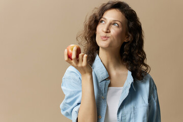 Wall Mural - Funny happy cute curly beautiful female in jeans casual shirt chewing enjoy apple posing isolated on over beige pastel background. Healthy food. Natural eco-friendly products concept. Copy space