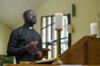 © pressmaster - African American priest in black casualwear holding rosary beads during pray while standing by pulpit with two burning candles