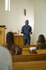 Wall Mural - Young black man in shirt with clerical collar and pants preaching in front of group of intercultural parishioners sitting on benches in church