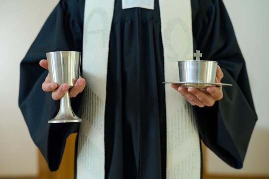 Hands of Catholic priest in cassock holding two cups with wine and unleavened bread for communion of parishioners of church