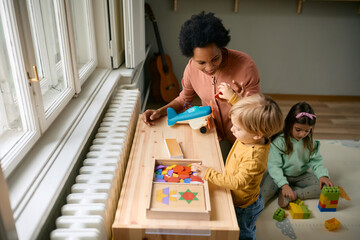 Wall Mural - Happy black teacher and small boy playing with puzzle shapes at kindergarten.