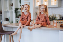 Two Scandinavian Cute Little Sisters With Ponytails Sitting On Kitchen Table Hugging Knees Looking At Camera. Little Swedish Girls In Pyjamas  On Breakfast Time At Home. Family Domestic Activities.