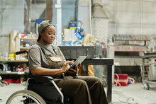 African Female Worker With Disability Using Digital Tablet At Work While Sitting In Wheelchair At Workshop