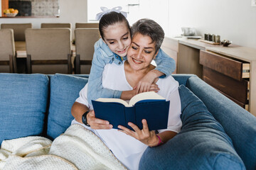 Wall Mural - hispanic Grandmother and granddaughter reading a book on sofa at home in Mexico Latin America