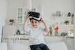 © Iona - Little cheerful boy smiling wide putting on vr glasses, happy to play using virtual reality googles sitting on sofa over blurry kitchen at home. Playfull Spanish boy at home using vr lenses for games.