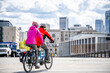 © vit - Two amateur cyclists wearing colorful jackets are cycling on a tandem bike through the streets of a modern city