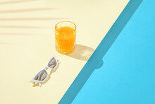 Minimal Tropical Composition With Stylish White Sunglasses And Orange Drink On Duo Tone Sandy And Blue Background. Creative Copy Space With Palm Leaf Shadow. Pool Party Concept.