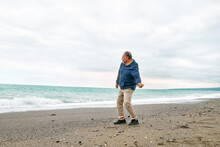 Happy Middle-aged Bearded Man Throws Pebbles Into The Sea, Walking Along Deserted Winter Beach In Cloudy Day. Concept Of Leisure Activities, Wellness, Freedom, Tourism, Healthy Lifestyle And Nature.