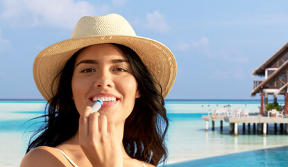 Wall Mural - beauty, cosmetics and summer holidays concept - portrait of happy smiling young woman in bikini swimsuit and straw hat applying lip balm over tropical beach background in french polynesia