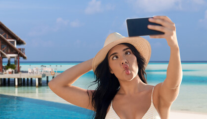 Wall Mural - travel, tourism and summer vacation concept - happy smiling young woman in bikini swimsuit and straw hat taking selfie with smartphone over tropical beach background in french polynesia