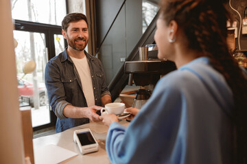 Female barista serving coffee at bar, giving cup to male customer. Cafeteria work. Rear. Back view.