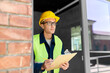 construction business and building concept - male builder in helmet and safety west with papers on clipboard and pencil looking out window