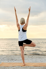 Wall Mural - fitness, sport, and healthy lifestyle concept - woman doing yoga tree pose on beach over sunset