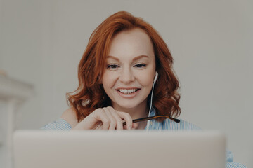 Pretty positive redhead woman gives online consultation, sits in front of opened laptop computer, has discussion with corporate client, smiles happily, uses modern earphones, enjoys online studying