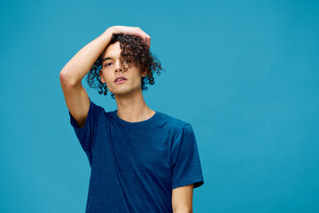 Handsome serious curly tanned Caucasian guy in basic t-shirt touches head posing isolated on over blue background. Lifestyle and Emotions concept. Good offer with copy space for ad