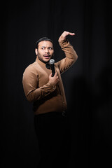 indian stand up comedian gesturing while telling joke into microphone on dark stage on black.