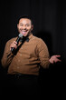 © LIGHTFIELD STUDIOS - joyful indian comedian in shirt and bow tie holding microphone during monologue on black.