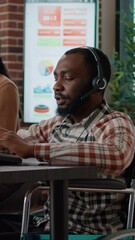 Wall Mural - Vertical video: African american man in wheelchair answering helpline call at customer service job, working in disability friendly office. Male worker using telework headset at helpdesk to help