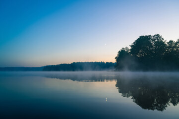 Beautiful mystical landscape. Forest lake at summer night before sunrise. Fog above calm water. Scenic nature.