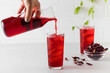 Herbal red tea made from hibiscus leaves is poured into a glass with ice from a jug. 