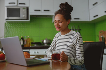 Wall Mural - beautiful smiling young woman holding cup of hot drink using laptop sitting at home