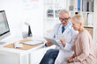 © Mediaphotos - Content senior male doctor with badge and stethoscope sitting at table and using tablet while explaining medical checkups result to mature woman