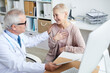 © Mediaphotos - Happy senior female patient with short hair sitting at table in doctors office and holding hand on chest while thanking doctor for good treatment and support