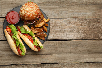 Tasty burger, hot dogs, potato wedges and sauce on wooden table, top view with space for text. Fast food