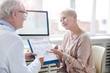 © Mediaphotos - Smiling senior female patient with blond hair sitting at table and pointing at paper in clipboard while asking doctor about prescribed drugs