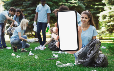 Wall Mural - Smiling Female Volunteer Showing Smartphone With Blank Screen While Collecting Garbage Outdoors