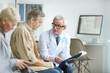 © Mediaphotos - Serious senior male doctor in white coat and eyeglasses holding clipboard with medical card and answering patients questions