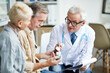 © Mediaphotos - Confident male doctor in white coat pointing at pill bottle and recommending medicine to senior couple