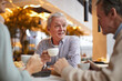 © Mediaphotos - Positive handsome gray-haired mature man in casual shirt sitting at table and talking to friends at tea party in cafe