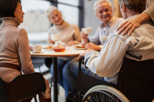 group of senior people visiting friend in wheelchair: they drinking tea and chatting at home